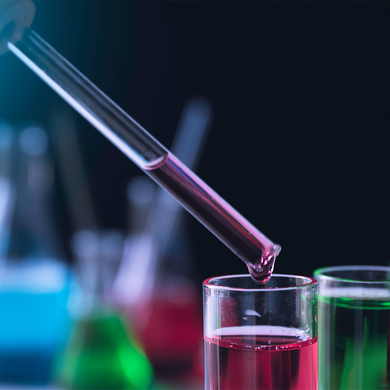 belis-loesung-chemisch Eine chemische rote Flüssigkeit wird von einem Glasröhrechen in ein anderes Glas getropft. Im Hintergrund stehen weitere bunte Flüssigkeiten in Glasbehältern.