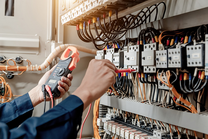 belis-oberflaechentechnik-branchen-elektrotechnik Jemand misst Strom an einer Wand mit elektrischen Bauteilen und Kabeln